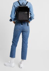 Czarna pikowana plecak z przednią kieszenią na zamek, regulowanymi paskami i małym uroczym detalem, noszony z dżinsowym strojem i białymi trampkami.