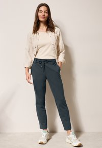 Chaqueta beige claro sobre una blusa crema, combinada con pantalones jogger gris oscuro y zapatillas claras. El look presenta un ajuste relajado y una cintura con cordón.