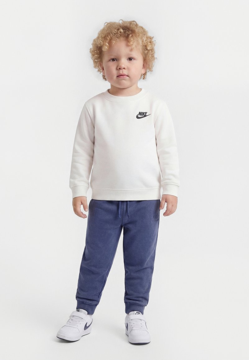 Νήπιο με σγουρά ξανθά μαλλιά που φοράει άσπρο φούτερ Nike, μπλε παντελόνι τζόγκερ και άσπρα αθλητικά παπούτσια Nike, στέκεται μπροστά σε απλό φόντο.