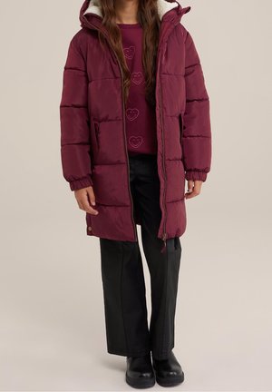 Personne portant un long manteau matelassé bordeaux avec capuche, un sweat bordeaux avec des cœurs souriants, un pantalon noir à jambes larges et des bottes noires.