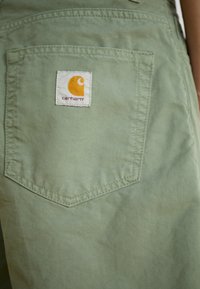 Green cotton fabric with a rear pocket featuring a white label and orange logo from Carhartt, showcasing a textured appearance and stitching details.