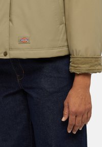 Giacca beige con una texture liscia, caratterizzata da un polsino a costine e una toppa con logo. Abbinata a jeans in denim scuro.