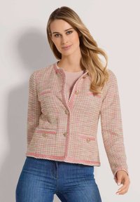 Tweedjacke in Pink mit strukturiertem Stoff, geflochtenem Besatz, goldenen Knöpfen und zwei Vordertaschen. Kombiniert mit einem hellpinkfarbenen Oberteil und Jeans.