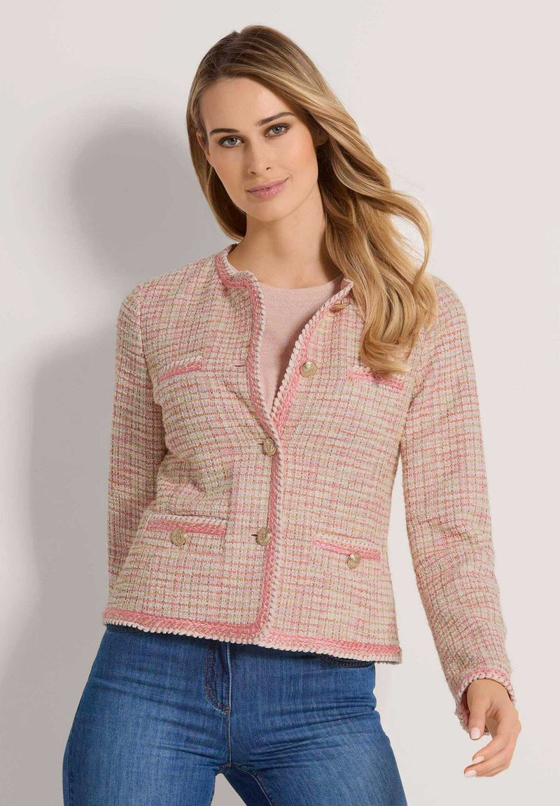 Tweedjacke in Pink mit strukturiertem Stoff, geflochtenem Besatz, goldenen Knöpfen und zwei Vordertaschen. Kombiniert mit einem hellpinkfarbenen Oberteil und Jeans.