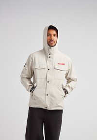 Lys beige vandtæt jakke med justerbar hætte, to brystlommer og sidelommer med lynlås. Sorte detaljer på manchetter og lynlås.