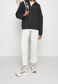 Černá bunda s předním zipem, elastickými manžetami a malým logem, v kombinaci s bílými kalhotami a béžovými teniskami. Pozadí je neutrální šedé.