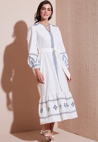 White long-sleeve dress with blue embroidery, tie belt at waist, tiered hem, and puffed sleeves. Worn with a pair of strappy sandals.