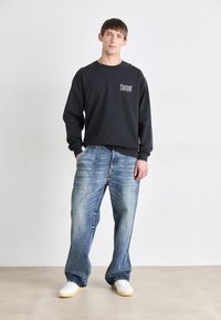 Schwarzer, langärmliger Sweatshirt mit dem Logo "Fantasy People", kombiniert mit hellblauen, abgewetzten weitem Jeans und weißen Turnschuhen.
