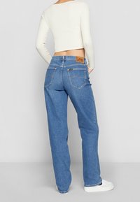 Modré džínové kalhoty s volným střihem, které mají zadní kapsy a opásání s béžovou nášivkou. Stylizované s přiléhavým dlouhým rukávem.