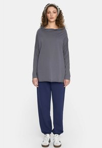 Kobieta z kręconymi włosami, ubrana w szary longsleeve, granatowe spodnie, białe trampki i szary opaskę na głowę, stojąca na tle jednolitej ściany.