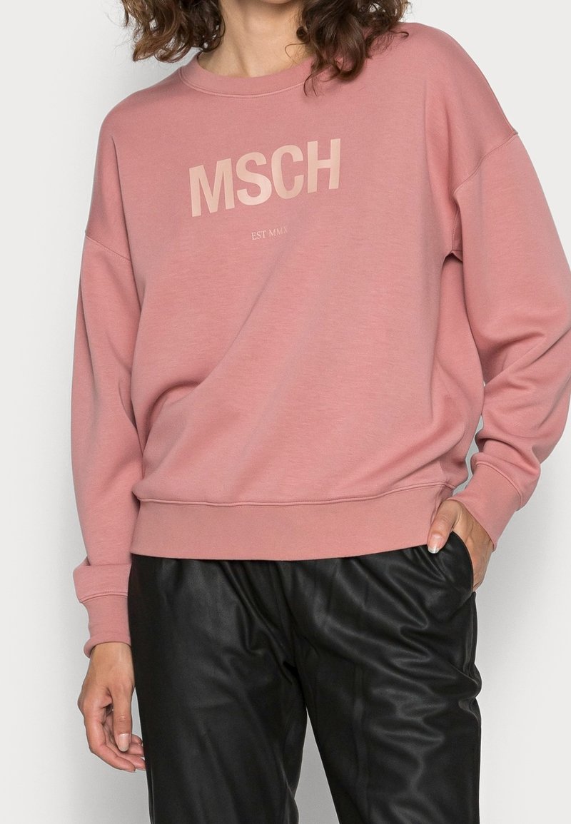 Femme portant un sweat-shirt rose poussiéreux avec le texte "MSCH" et un pantalon noir, debout avec une main dans la poche et les cheveux bouclés partiellement visibles.