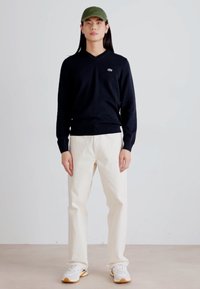 Námornícky sveter s V-výstrihom, svetlo béžové nohavice a biele tenisky. Zelená čiapka dotvára vzhľad. Uvoľnený strih s jednoduchým dizajnom.