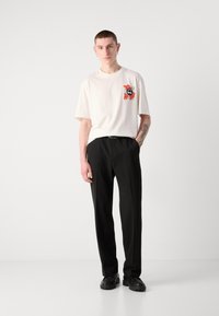 Šviesiai ruda trumpa rankovė t-shirt su juodu ir oranžiniu piešiniu ant krūtinės, derinama su juodomis pritaikytomis kelnėmis ir juodais masyviais batais.