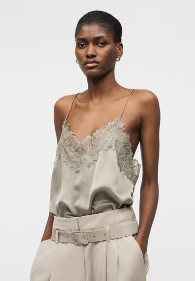 Femeie care poartă o camisola din satin bej, cu dantelă, cu bretele subțiri, băgată în pantaloni cu talie înaltă cu curea, mâinile în buzunare.