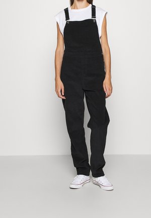Woman wearing black corduroy overalls, white sleeveless shirt, and white sneakers standing against a plain light background.