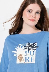 Blaues Langarmhemd mit floralem Druck in Weiß, Braun und Marine, mit dem Wort "PURE" in fetten Buchstaben. Glatte Stofftextur.