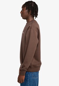 Un joven con el cabello trenzado se encuentra de lado, vistiendo un sudadera marrón y unos vaqueros de denim azul, contra un fondo blanco liso.