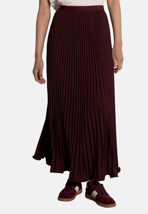 Femme portant une longue jupe plissée bordeaux foncé assortie à des baskets bordeaux avec des bandes blanches.