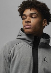 Felpa grigia con zip, dotata di una cerniera nera e un logo Jordan nero sul petto. Realizzata in un tessuto morbido con una finitura testurizzata.