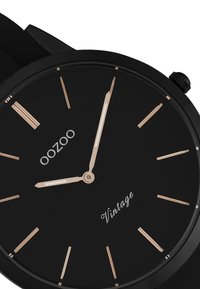 Montre analogique noire au design minimaliste. Dispose d'un cadran noir, d'index et aiguilles en or rose, avec l'inscription "OOZOO Vintage".