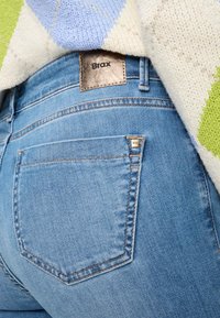 Jean en denim bleu clair avec une étiquette en cuir "Brax" sur la ceinture et une petite étiquette logo sur la poche arrière, affichant un design classique.