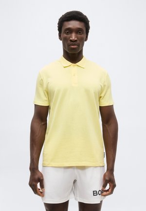 Muškarac nosi blijedožutu polo košulju i bijele kratke hlače s logotipom "BOSS", stoji ispred jednobojne svijetle pozadine.