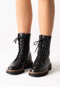 Botas de combate de cuero negro con una suela robusta, cordones en la parte delantera, cremalleras en los laterales y detalles en marrón cosidos en el borde de la suela.