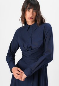 Mörkblå, långärmad skjortklänning med knapplist framtill, krage och knutdetalj i midjan, tillverkad av ett slätt tyg.