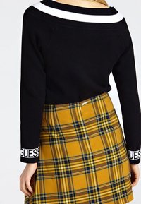 Schwarzer, schulterfreier Pullover mit weißen Streifen und langen Ärmeln, kombiniert mit einem gelben Karorock, der blaue und braune Linien aufweist und einen seitlichen Reißverschluss hat.