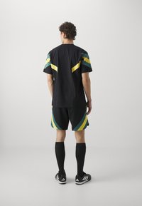 Camiseta deportiva negra con rayas verdes y amarillas, combinada con pantalones cortos negros que tienen acentos similares. Calcetas altas negras y zapatillas deportivas.