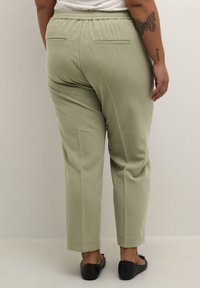 Pantalon vert olive clair à coupe décontractée, avec une taille élastique et deux poches arrière. Porté avec des chaussures plates noires.