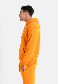 Sudadera con capucha naranja hecha de tela suave, con un bolsillo tipo canguro, cordón ajustable y puños acanalados, combinada con pantalones a juego.