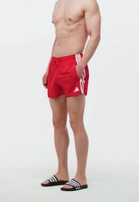 Rote Schwimmshorts mit weißen Seitenstreifen und elastischem Bund. Getragen mit schwarzen Rutschen mit weißen Streifen.