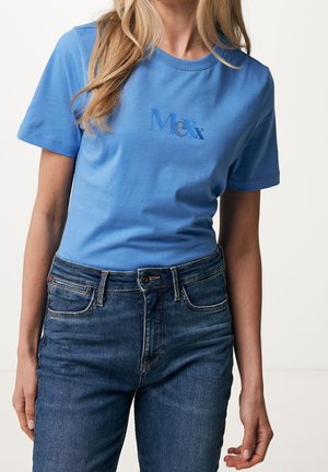 Frau trägt ein hellblaues T-Shirt mit dem Text "Mex" und hoch taillierte blaue Jeans, steht vor schlichtem hellem Hintergrund.