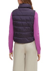 Gestepptes lila Weste mit hohem Kragen, horizontalen Nähten und lockerer Passform, getragen über einem rosa Langarmshirt und cremefarbenen Hosen.