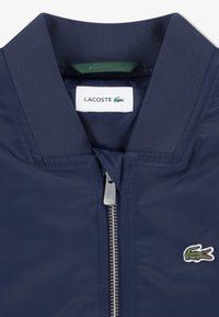 Veste bleu marine avec une fermeture éclair, un col côtelé et un accent vert. Logo Lacoste brodé sur le côté gauche de la poitrine. Texture lisse.