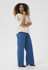 Chemise beige texturée avec broderies florales sur les manches, associée à un jean en denim bleu ample et des chaussures marron foncé. Pose debout.