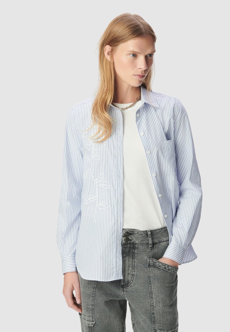 Camicia a righe blu e bianche con maniche lunghe, caratterizzata da un design floreale ricamato sul lato sinistro. Abbinata a una maglietta bianca.