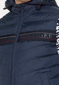 Marineblaues Steppweste mit einem Reißverschluss vorne, versehen mit horizontaler Naht, einem Logo-Bund in Schwarz mit roten und weißen Streifen sowie einer gemusterten Kapuze.