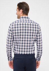 Man die wegkijkt en een langemouwen shirt met een navy, witte en lichtbruin geruit patroon draagt, ingestopt in donkerblauwe broeken met knoopzakken aan de achterkant.