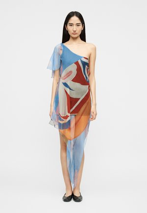 Kvinde iført en asymmetrisk kjole med en skulder og abstrakte blå, røde, hvide og orange mønstre samt sorte flade sko, stående mod en ensfarvet baggrund.