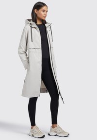Lichtgrijze lange jas met een capuchon, ritssluiting aan de voorkant, zijzakken en zwarte trekkoorden, gecombineerd met zwarte leggings en beige sneakers.