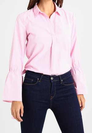 Kobieta w różowej bluzce z długimi rękawami zakończonymi rozszerzanymi mankietami oraz ciemnoniebieskich jeansach z wysokim stanem na białym tle.