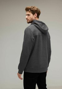 Sudadera gris con un panel texturizado en la parte posterior, que cuenta con capucha ajustable y puños acanalados. El material parece suave y cómodo.