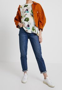 Orange Jacke, weißes, gemustertes Hemd mit buntem Gemüse, blaue Jeans und weiße Sneaker. Die Jacke hat einen lässigen Schnitt und Knöpfe.
