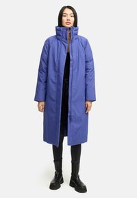 Woman wearing long, purple padded coat with high collar, black top, black skinny pants, and black chunky ankle boots, standing on white background.