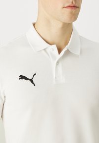 Weißes Poloshirt aus strukturiertem Stoff, zwei Knöpfe am Kragen und ein schwarzes springendes Puma-Logo auf der linken Brust.