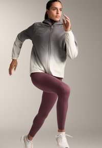 Ľahká, gradient šedá bunda s kapucňou, so zipsom a elastickými manžetami, kombinovaná s priliehavými vínovými legínami a bielymi športovými topánkami.