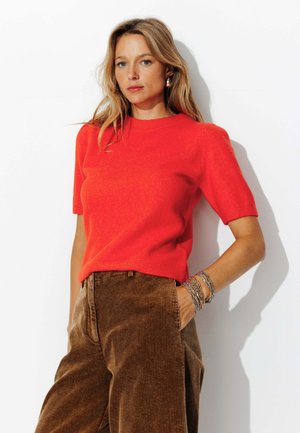 Pull rouge à manches courtes avec un col rond, fabriqué à partir d'un matériau texturé. Associé à un pantalon large en velours côtelé marron.