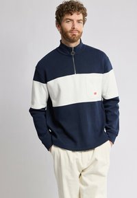 Pull à col haut, zippé quart et poche frontale, en bleu marine et blanc avec un design coloré. Tissu doux, coupe décontractée, accents de style minimalistes.